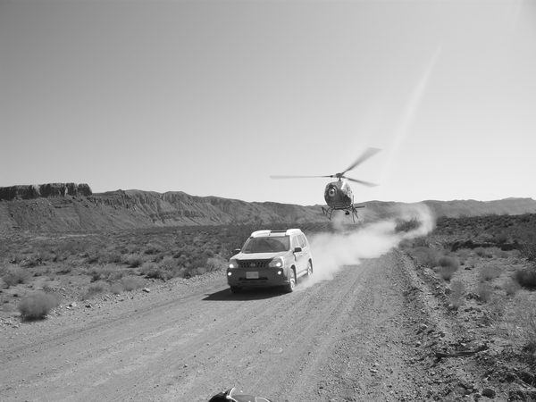 Black and white photo of helicopter flying low over dirt road behind SUV, Tenerife